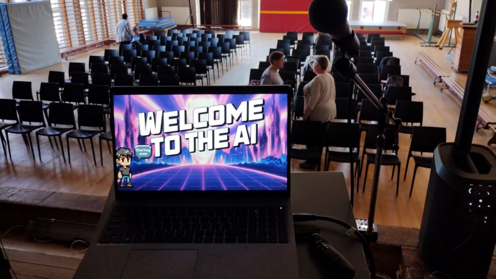 Een laptop met het scherm "Welcome to the AI" staat op een podium in een lege zaal met stoelen, met een microfoon op de voorgrond en enkele mensen die in de achtergrond praten. 